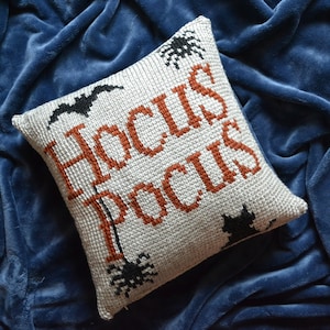 Pode incluir: Almofada quadrada, branco sujo, com as palavras "Hocus Pocus" em laranja e preto. A almofada apresenta um desenho de morcego, aranhas e gato. A almofada está sobre um cobertor azul.