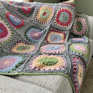 May include: A crocheted throw blanket with a gray background and colorful circular patterns. The blanket is made of yarn in shades of pink, blue, green, and yellow.