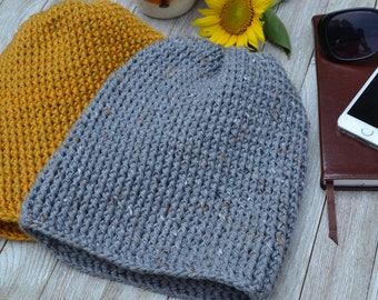 May include: Two crocheted beanies, one in a mustard yellow color and the other in a speckled gray color. The beanies are sitting on a wooden surface.