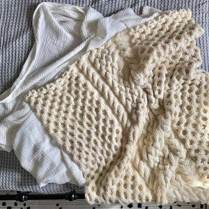 May include: A white crocheted throw blanket with a cable knit pattern. The blanket is folded and draped over a white and gray textured surface.