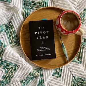 May include: A wooden tray with a black book titled "The Pivot Year: 365 Days to Become the Person You Truly Want to Be" by Brianna Wiest, a red and white polka dot mug with ice and a brown liquid, and a green and white crochet blanket.