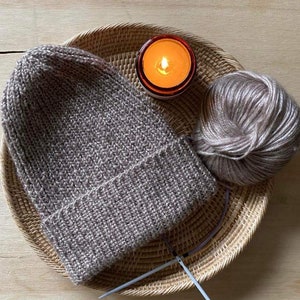 Könnte beinhalten: Eine handgefertigte graue Strickmütze mit umgeschlagenem Saum liegt auf einem geflochtenen Korb. Ein Knäuel Wolle und Stricknadeln liegen neben der Mütze. Eine brennende Kerze in einem Glas erzeugt ein warmes Leuchten.