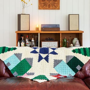 Puede incluir: Una manta de ganchillo colorida con patrones geométricos en tonos de verde, azul y blanco. La manta está colocada sobre un sofá de cuero marrón.