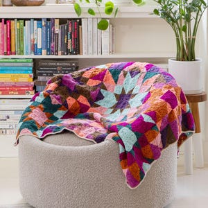 May include: A colorful crocheted blanket with a star pattern in shades of pink, purple, teal, brown, and orange. The blanket is draped over a light gray, round ottoman.