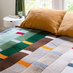 Peut inclure: Une couverture en tricot multicolore avec des rayures vertes, brunes, orange, rouges et bleues. La couverture est pliée sur un lit avec deux oreillers.