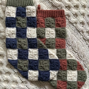 May include: Two handmade Christmas stockings. One is mainly navy blue, olive green, and cream, the other rust, olive green, and cream. Both have a checkerboard pattern and are made of yarn.