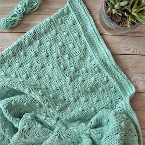 Puede incluir: Una manta de punto suave de color verde menta con un patrón de puntadas texturizadas. La manta está doblada y drapeada sobre una superficie de madera.
