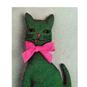 Puede incluir: Un gato de fieltro verde con una pajarita rosa. El gato está sentado con la cola enroscada alrededor de sus patas.