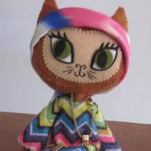 May include: A handmade felt cat doll wearing a colorful zig-zag patterned dress and a pink and blue tie-dye headband. The doll has large green eyes and a gold chain necklace.
