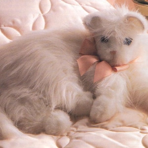 May include: A plush white cat toy with a pink bow around its neck, resting on a quilted, light pink surface. The cat has blue eyes and a fluffy appearance, creating a soft and cuddly aesthetic. The background is a matching light pink, enhancing the toy's gentle appeal.