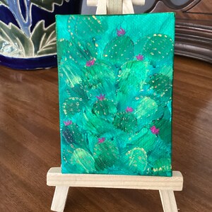 May include: A small painting on canvas depicting a cluster of green cactus plants with pink flowers. The painting is on a small easel.