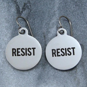 May include: A pair of silver earrings with round discs that say "RESIST" in black lettering.