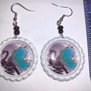 May include: A pair of white bottle cap earrings with a black dragon with blue wings design. The earrings are about 1 inch in diameter.