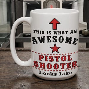 May include: A white ceramic mug featuring a red arrow pointing upward, a red star, and black and red text that reads: THIS IS WHAT AN AWESOME PISTOL SHOOTER Looks Like, surrounded by decorative red stars on a white background.