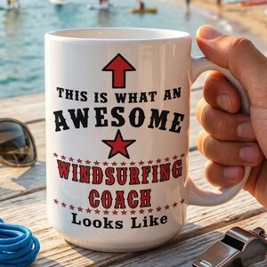 May include: A white ceramic mug with a red arrow pointing up, a red star, and black and red text that reads: This is what an awesome windsurfing coach looks like. The mug is positioned on a wooden surface next to sunglasses, blue cord, and a metal whistle.