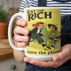 May include: White ceramic mug with a large handle. The mug features the text "EAT THE RICH" above an illustration of two cartoon figures. Below the illustration is the text "save the planet". The mug is held by a person.