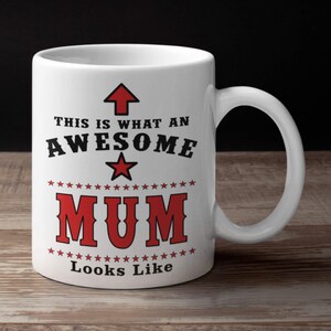 May include: White ceramic mug with a curved handle. The mug features the text "THIS IS WHAT AN AWESOME MUM LOOKS LIKE" in black and red lettering, with a red star and arrow design.