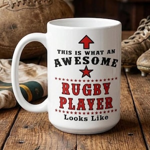 May include: White ceramic mug with a large handle, featuring the text "THIS IS WHAT AN AWESOME RUGBY PLAYER LOOKS LIKE" in black and red. An upward-pointing red arrow and a red star are above the text. The mug is on a wooden surface.