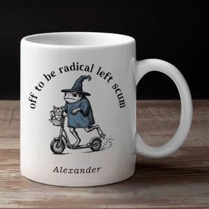 May include: White ceramic mug featuring a black illustration of a frog in a wizard hat riding a scooter, with a cat in a basket. The mug has the text "off to be radical left scum" and "Alexander".