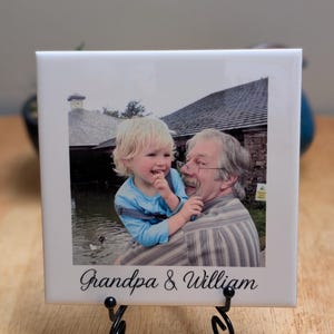 May include: A white ceramic tile featuring a photograph of a child and an older adult. The child is wearing a blue shirt and overalls, and the adult is wearing a striped shirt. The tile is displayed on a black metal stand, with the words "Grandpa & William" in script.