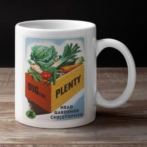 May include: White ceramic mug featuring a vintage illustration of a wooden crate overflowing with fresh vegetables. The crate is labeled "DIG FOR PLENTY" with "Head Gardener Christopher" below. The mug has a curved handle.