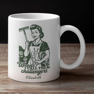 May include: White ceramic mug featuring a vintage-style illustration of a woman holding a rake and a snail. The mug has the text "Weeds? I call them challengers" and the name "Elizabeth" in green.