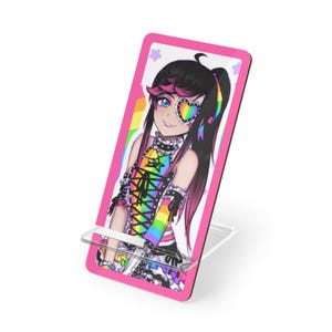 May include: A rectangular phone stand featuring a vibrant illustration of an anime-style character with dark hair, a rainbow-coloured outfit, and a heart-shaped rainbow eye patch. The stand has a bright pink border and a clear acrylic support base.