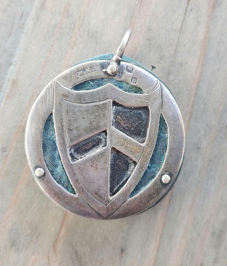 Pu&ograve; includere: Ciondolo in argento con disegno a scudo. Il ciondolo presenta una forma a scudo al centro, circondata da una cornice circolare. Lo scudo ha una superficie strutturata con aree scure. Il ciondolo ha un anello nella parte superiore per appenderlo.