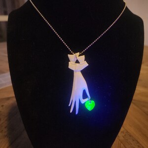 May include: A white ceramic necklace with a hand holding a green heart pendant. The pendant glows under a black light.