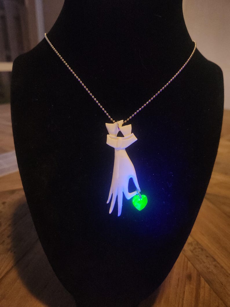 May include: A white ceramic necklace with a hand holding a green heart pendant. The pendant glows under a black light.
