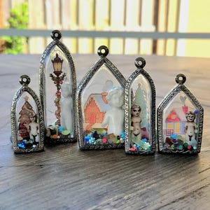 Puede incluir: Una colección de cinco colgantes en miniatura con temática navideña. Cada colgante presenta una pequeña figura dentro de una carcasa de vidrio transparente en forma de arco con un marco decorativo de color plateado. Los colgantes contienen árboles de Navidad, casas y figuras en miniatura.