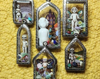 May include: A collection of six silver-toned pendants, each containing a miniature figurine and colorful stones. The pendants feature various themes, including a fairy, a skull, and a doll, set against a yellow background.