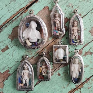 Puede incluir: Siete colgantes de plata con frentes de vidrio, cada uno contiene una pequeña figurita de porcelana. Las figuritas son de varias poses y estilos, algunas con elementos decorativos adicionales dentro del vidrio.
