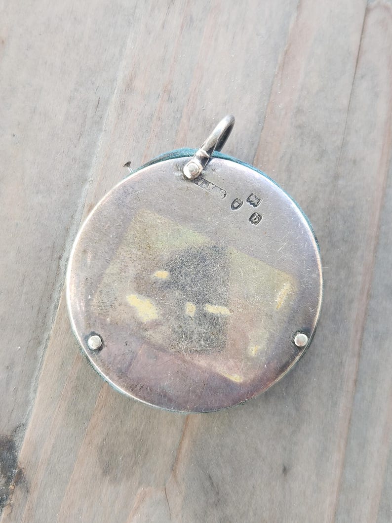 Pu&ograve; includere: Un ciondolo rotondo in argento con un bordo in tessuto turchese. Presenta un piccolo anello nella parte superiore per una catena. Il retro del ciondolo presenta marchi e una piccola finestra rettangolare.