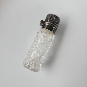 May include: A clear, faceted glass perfume bottle with a silver-colored metal cap. The cap has a floral design and a hinged closure. The bottle's body is a series of diamond-shaped facets.
