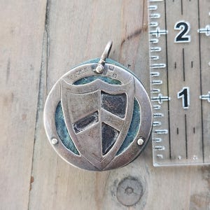 Pu&ograve; includere: Ciondolo argentato con un design a scudo, con un motivo a scudo centrale e una cornice circolare. Il ciondolo misura circa 3,8 cm di diametro e pende da un piccolo anello. Lo sfondo &egrave; un tessuto verde acqua.