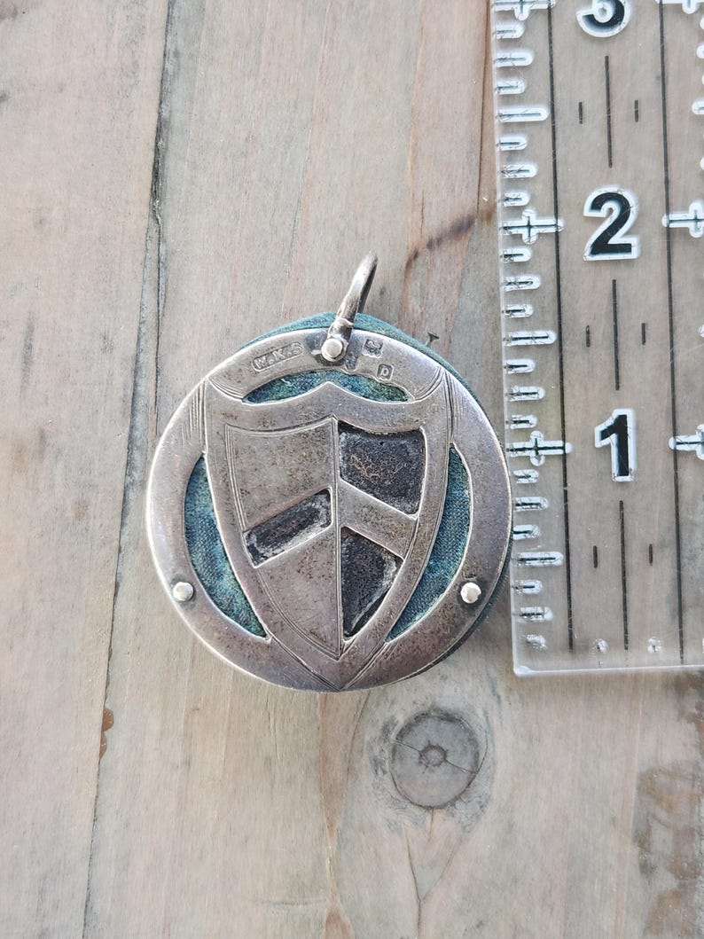 Pu&ograve; includere: Ciondolo argentato con un design a scudo, con un motivo a scudo centrale e una cornice circolare. Il ciondolo misura circa 3,8 cm di diametro e pende da un piccolo anello. Lo sfondo &egrave; un tessuto verde acqua.