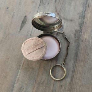 May include: Open silver compact with a mirror, containing pink powder and a matching powder puff. The compact has a chain and a ring. The puff is light pink and round.