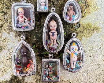 May include: A collection of seven silver-toned metal pendants, each featuring a clear casing filled with small colorful stones and a miniature figurine. The figurines include various characters such as a clown, a dark-skinned figure, and babies.