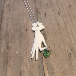 May include: A white ceramic hand pendant necklace with a green glass heart charm. The hand is holding the heart charm.