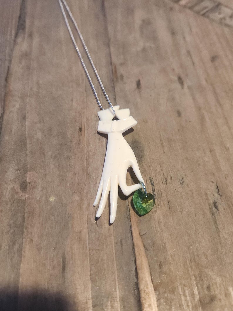 May include: A white ceramic hand pendant necklace with a green glass heart charm. The hand is holding the heart charm.
