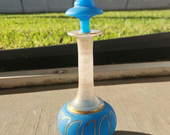 May include: A blue glass bottle with a gold trim and a blue glass stopper. The bottle has a round body and a long, thin neck.