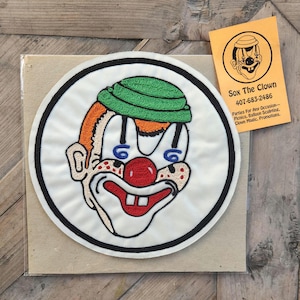 Könnte beinhalten: Gestickter Aufnäher eines Clowns mit grünem Hut, roter Nase und einem breiten Lächeln. Der Aufnäher ist weiß mit schwarzem Rand und rund. Der Text "Sox The Clown" und eine Telefonnummer sind im Hintergrund sichtbar.