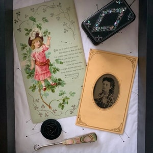 May include: A framed shadow box with a vintage postcard featuring a young girl in a pink dress and a white hat, a black and white photograph of a woman, a black button, a small black and iridescent box, and a crochet hook with a colorful handle.