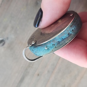 Pu&ograve; includere: Un cuscino per spilli ovale color argento con un bordo in tessuto blu. Il cuscino per spilli ha un piccolo anello di metallo curvo attaccato. Il tessuto mostra segni di usura e invecchiamento, con qualche scolorimento.