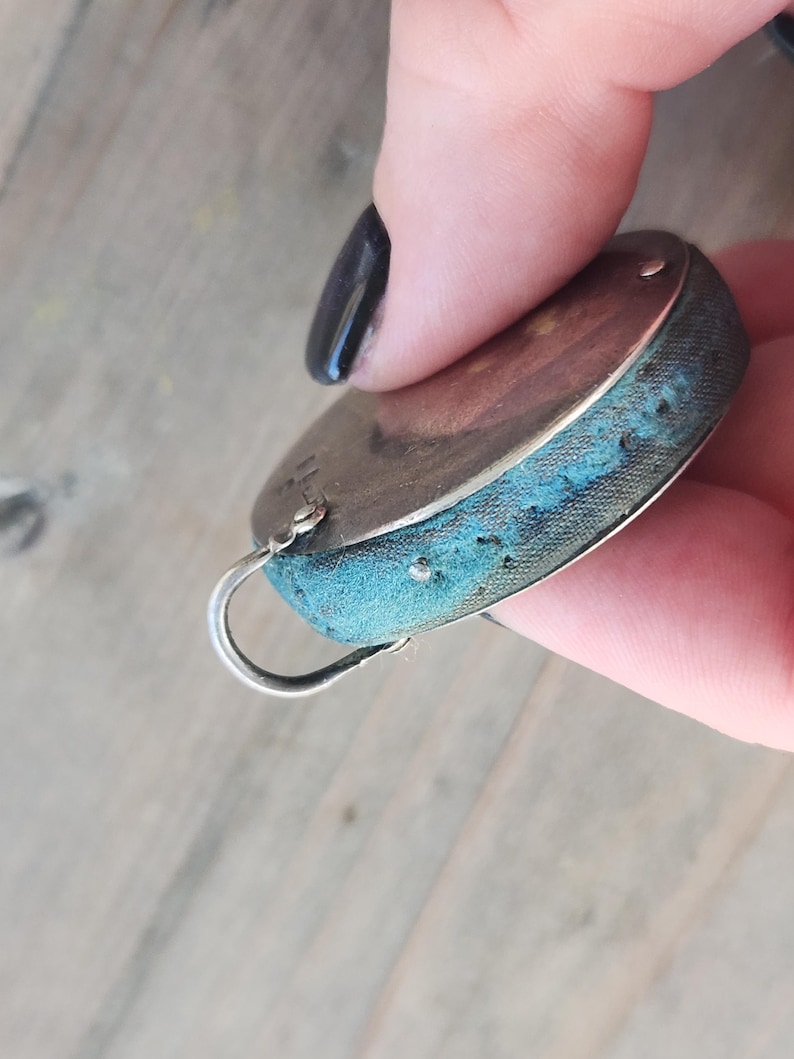 Pu&ograve; includere: Un cuscino per spilli ovale color argento con un bordo in tessuto blu. Il cuscino per spilli ha un piccolo anello di metallo curvo attaccato. Il tessuto mostra segni di usura e invecchiamento, con qualche scolorimento.