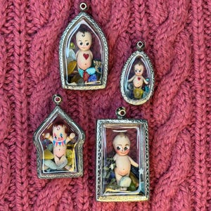 Op de afbeelding: Vier vintage-stijl hangers met kleine poppenfiguren in heldere rechthoekige en ovale frames. De poppen hebben geschilderde gezichten en zijn omgeven door kleurrijke stenen. De frames zijn zilverkleurig en hebben een lus aan de bovenkant om op te hangen.