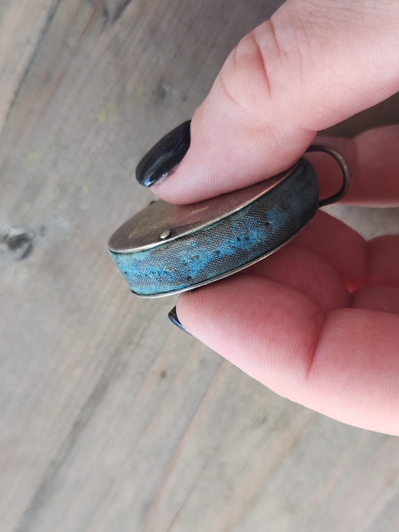 Pu&ograve; includere: Un piccolo cuscino per spilli rotondo antico con una cornice in metallo color argento e un cuscino in tessuto blu. Il cuscino ha un motivo cucito decorativo. Un piccolo anello di metallo &egrave; attaccato in alto.