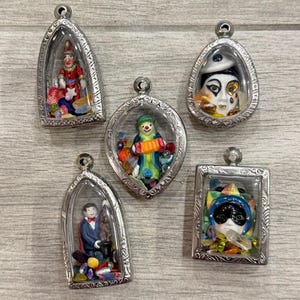 May include: Five silver-toned metal pendants with clear glass fronts, each containing a miniature figurine and colourful beads. The figures include a jester, a clown with an accordion, a masked face, a magician with a rabbit, and a colourful harlequin mask.