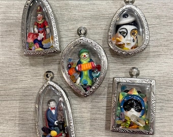 May include: Five silver-toned metal pendants with clear glass fronts, each containing a miniature figurine and colorful beads. The figures include a jester, a clown with an accordion, a masked face, a magician with a rabbit, and a colorful harlequin mask.